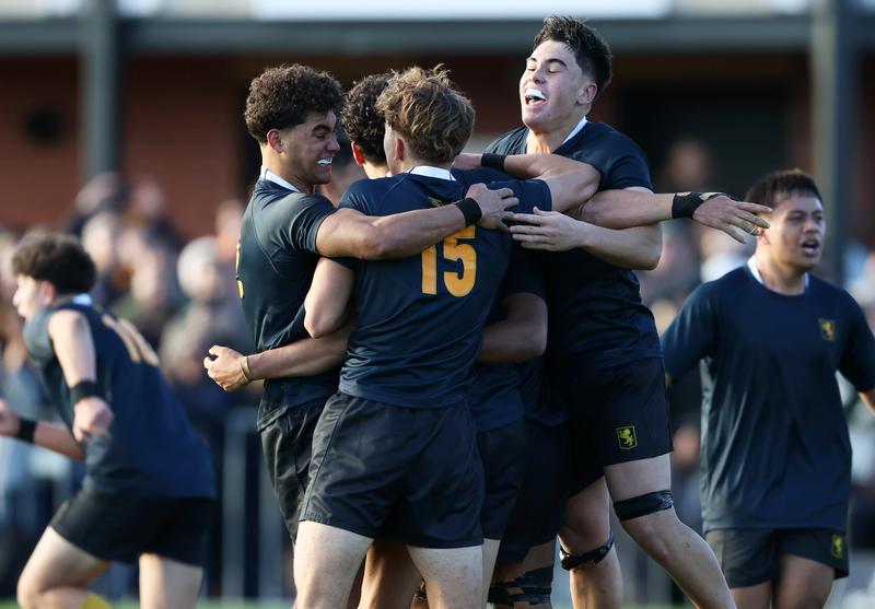 A group of young male rugby players, jumping and celebrating smiling and looking tired from playing, with people watching the game from the background